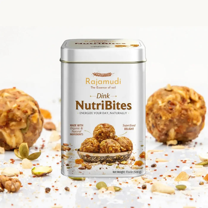 A tin of Rajamudi Dink NutriBites energy bites, with the product name and benefits listed on the label, surrounded by dry fruits such as almonds and seeds, and individual bites visible in the foreground.